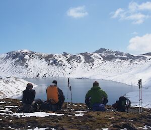 During field training — Jaimie Marty and Kris, sitting on the grass with their backs to the camera, having lunch while they take in the view of Waterfall Lake. Thick snow cover on the lakes shore and the hills in the background