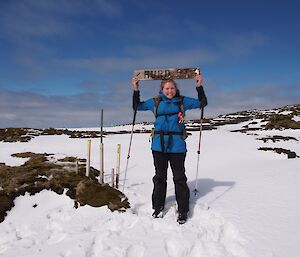 Jaimie, standing on a thick snow covered track, showing the way to Hurd Point, by holding the wooden Hurd Point sign over her head