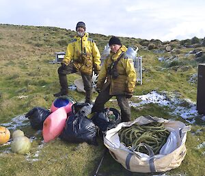 Chris and Clive standing on the grass at Bauer Bay hut with the bags of marine debris and various fishing floats, including the first load of rope packed in a bulker bag