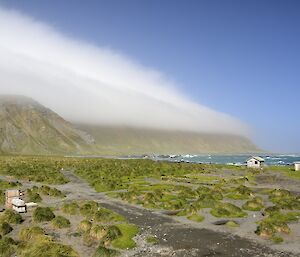 Orographic cloud over the island. Two layers of cloud can be seen capping the escarpment, while in the foreground is the tussock covered isthmus and the huts of the magnetic quiet zone
