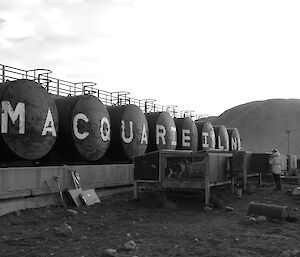 A black and white photo of the dog kennels in front of the fuel farm. The fuel tanks have the big letters painted on them spelling MACQUARIE ISLAND