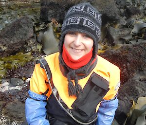 Bianca sampling juvenile sea stars from the rock pools of Garden Cove. She is holding a rock on which you can see a few small orange sea stars. She is wearing a dry suit and has blue, rubber gloves which extend up her arms just past her elbows