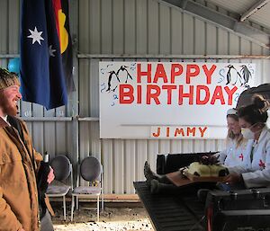 In the boat shed — ‘Nurses’ Jaimie and Laura presenting Jimmy with his birthday cake. On the wall in the background is a large sign saying "Happy Birthday Jimmy". The Australian and the Aboriginal flags are hanging from the ceiling.