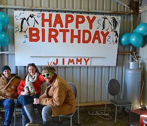 In the boat shed on Australia Day celebrating Jimmy’s birthday. Jimmy, wearing green heart shaped sunglasses, is sitting with Jess and Robbie. They are sitting to the left of and below a large sign that has "Happy Birthday Jimmy" written on it. There are a bunch of blue balloons on either side of the sign