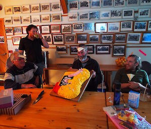 Celebrating Greg Bird’s birthday. Greg is sitting at one of the dining tables with his cake. Three other people are around him. Each is wearing either a paper beak or feathers in their hair