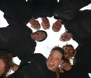 New Zealand Team bonding session, with an itinerant Aussie photo bomber. Eight members of the team, dressed in black, are standing in a circle. The view is from below of their faces. In the bottom left corner part of the face of one of the Aussie team members wanting to spoil their session