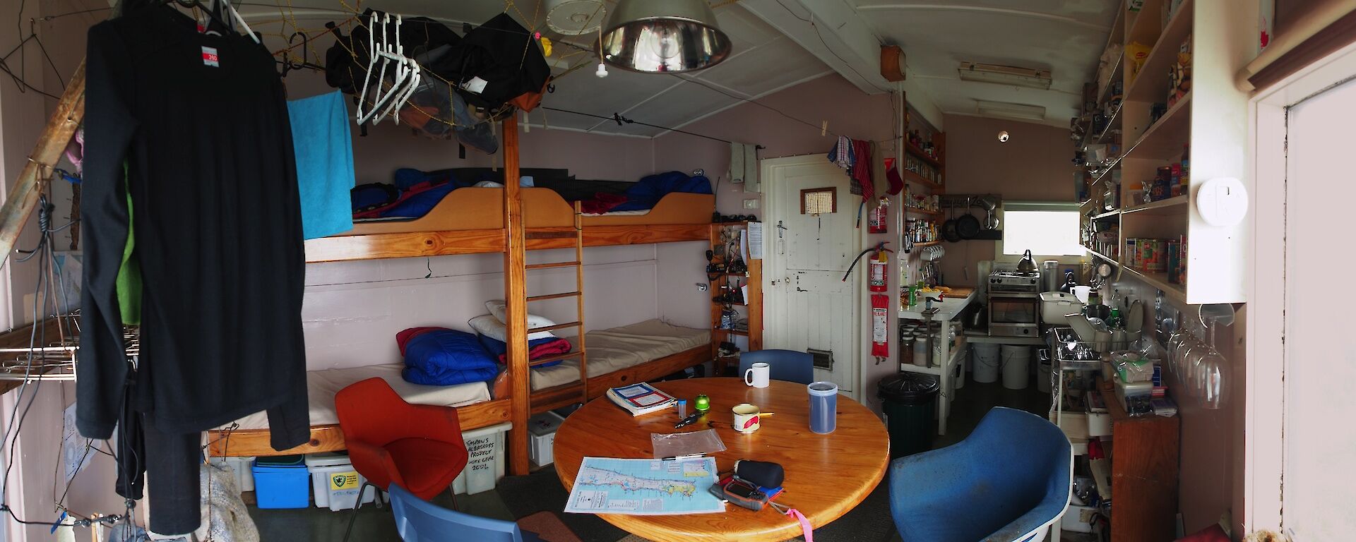 My home while down south — Hurd Point hut. A panoramic view of the inside of the hut, showing from the left, clothes hanging to dry, two double bunks on the back wall, table and chairs then through to the kitchen area and shelves stacked with food (on the far right)