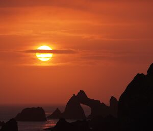 A stunning sunset viewed from Hurd Point hut showing the orange orb of the sun filtering through the cloud and the rugged offshore rock stacks (including an arch) silhouetted