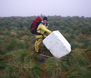 Barry with the styrofoam block in July 2013. Barry, dressed in his yellow wet wether gear and carrying his survival backpack, with the block leaning on an angle. They are amongst the tussock grass. The block measures 100cm by 100cm by 62cm