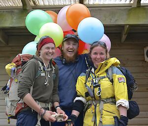 Ange and Leona, both wearing there wet weather gear and carryon back packs, arrive back at station just as Aaron brings balloons to decorate the mess for Jaimie’s birthday. They are standing on the cold verandah with the multi coloured balloons around their heads