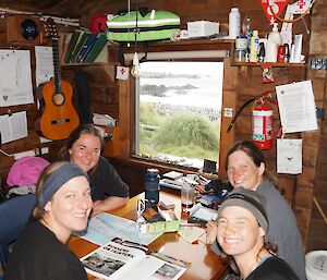 Green Gorge Birthday Breakfast. Leona, Dana, Ange and Karen sit around the table at Green Gorge hut. Throught he window behind the group you can see the tussock and many penguins on the beach