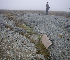 Bianca and Keith standing at the top end of the trench. There is mist drifting over the site. A metal panel lies half buried about halfway along the trench and a rusty pipe can be seen exposed near the top of the trench