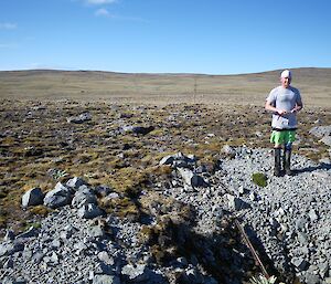Josh standing next to the trench — that lines up with the Island Lake Track which can be seen in the distant background. It is a clear sunny day and Josh is wearing shorts, T-shirt, though he is wearing gumboots. A rusted star picket can be seen in the end of the trench