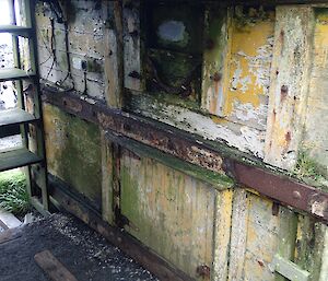 Cold porch of the Sandy Bay hut. There is a rusted steel cross-member and the panels of the wall are covered in mould, moss and cracked, fading dull yellow paint. The floor is covered in dirt and feathers from the moulting penguins