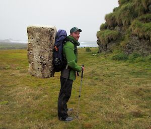 Kris, standing side on somewhere on the west coast with a block of styrofoam strapped to the back of his pack. He is staining on the featherbed beside a large rock stack. The block of foam is half his height and approximately 40cm wide