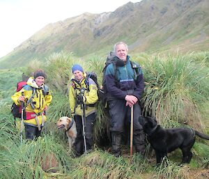 The happy search team of Kate, Karen Nick and the two dogs — Finn (golden labrador) and Waggs (black labrador). They are sitting amongst the tussock with the rugged slopes of the escarpment in the background