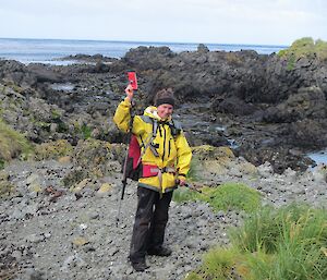 The notebook is found! Kate dressed in her yellow wet weather gear, holds up the red notebook. She is standing on the rocky coast with the ocean in the background