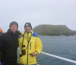 Two expeditioners smiling on the ship with Macquarie Island in background