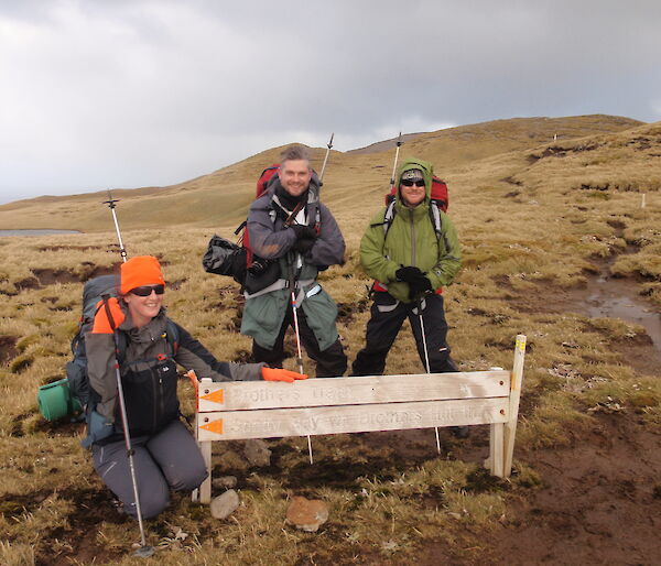 Three expeditioners pose beside a walking track sign