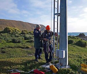 Two expeditioners check the safety harness of one of them, beside the mast