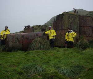 Expeditioners stand amongst large rusted barrels known as penguin digesters, surrounded by lush green grasses