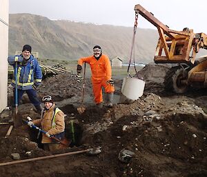 One expeditioner in a ditch with two standing on ground level with shovels, a small excavator sits to their right