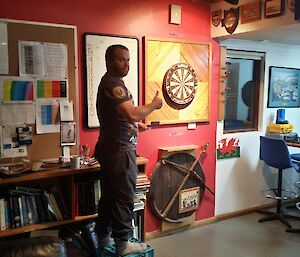 Greg stands on a milk crate next to the dart board