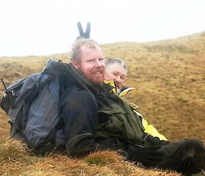 Two expeditioners sit on the ground, one doing rabbit ears with his fingers behind the other guy’s head