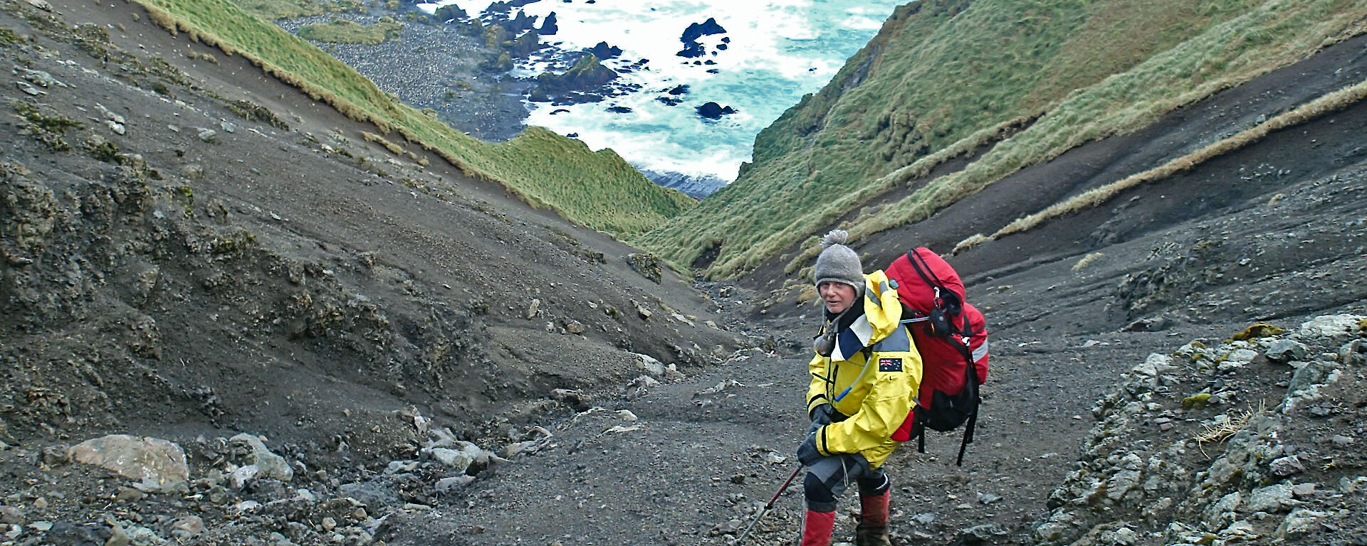 Expeditioner on a steep slope covered in soil but flanked by moss, looks back at the camera with the ocean and rocks behind him