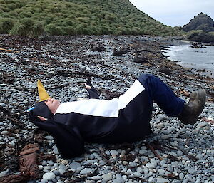 A man in a penguin costume lies on his back on the rocky shore