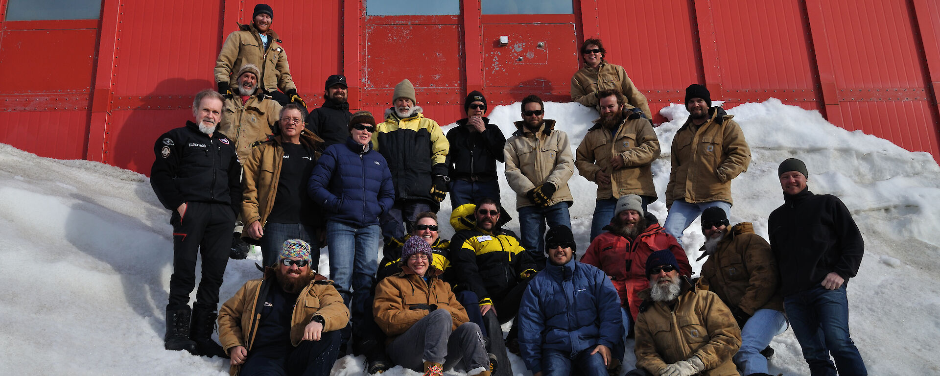 Twenty one people standing and sitting on the snow down wind of the red living quarters
