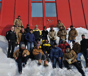 Twenty one people standing and sitting on the snow down wind of the red living quarters