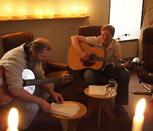 Two expeditioners sitting in seats playing guitars for the carol singing and behind them numerous candles