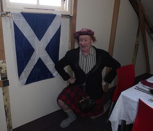 An expeditioner wearing a kilt kneeling next to the Scottish flag