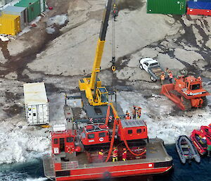 A photo taken from a helicopter looking down on a crane lifting a Hägglunds all terrain vehicle from a jet barge