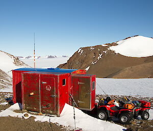 Small and red Fang Hut with two quad bikes parked just outside and snowy peaks in the background