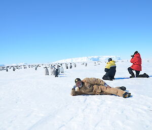 Geoff Brealey lies on snow with many emperor pnguins in the background