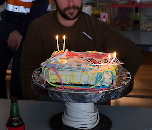 Expeditioner poses with electrician themed birthday cake featuring warning sticker and words written out in electrical cords made of icing