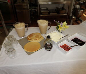 A table covered with white linen tablecloth shows morning tea items including pancakes, maple syrup and more