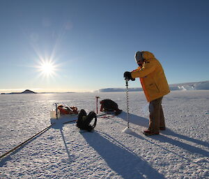 Justin drilling sea ice with handheld drill and sun shining in background