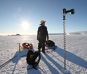 Drilling ice off Mawson an expeditioner stands surrounded by equipment with sun shining in background