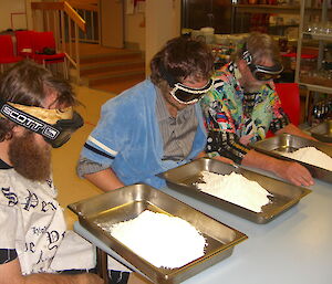 Three expeditioners wear goggles and face metal tubs filled with flour for a game on quiz night