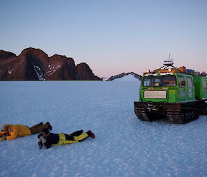 Two expeditioners on their bellies on the ice taking photos of a sunset with a Hägglunds overland vehicle in the background