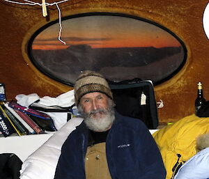 Geoff Brealey poses inside Beche hut with an orange glow to the horizon seen through a window behind him