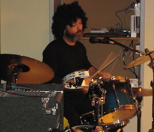 Expeditioner playing drums and wearing a very curly wig.