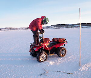 An expeditioner standing on the front of a quad bike to drill into the ice.