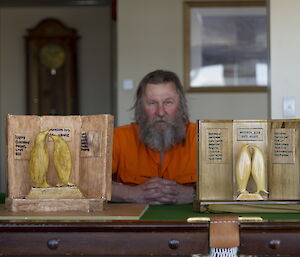 Man sits between his wooden wall plaque and the cake that is decorated to look exactly the same