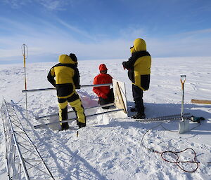Three people in Antarctic warm gear attaching a metal plate to the rodsto weather station install