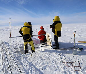 Three people in Antarctic warm gear attaching a metal plate to the rodsto weather station install