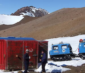Two male expeditioners stand outside Fang hut and next to a Hägglunds vehicle